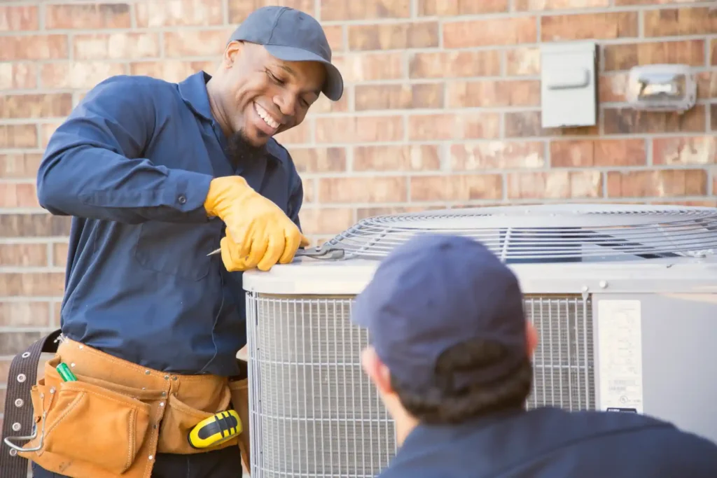HVAC technicians in blue uniforms servicing an outdoor air conditioning unit near a brick wall with electrical meter and conduit