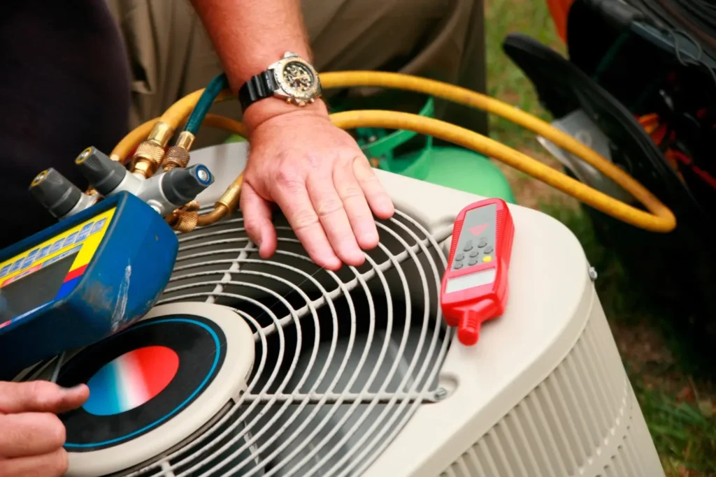 A close-up shows a person's hands working with gauges and a thermometer on top of an outdoor air conditioning unit. Yellow and green hoses are also visible.