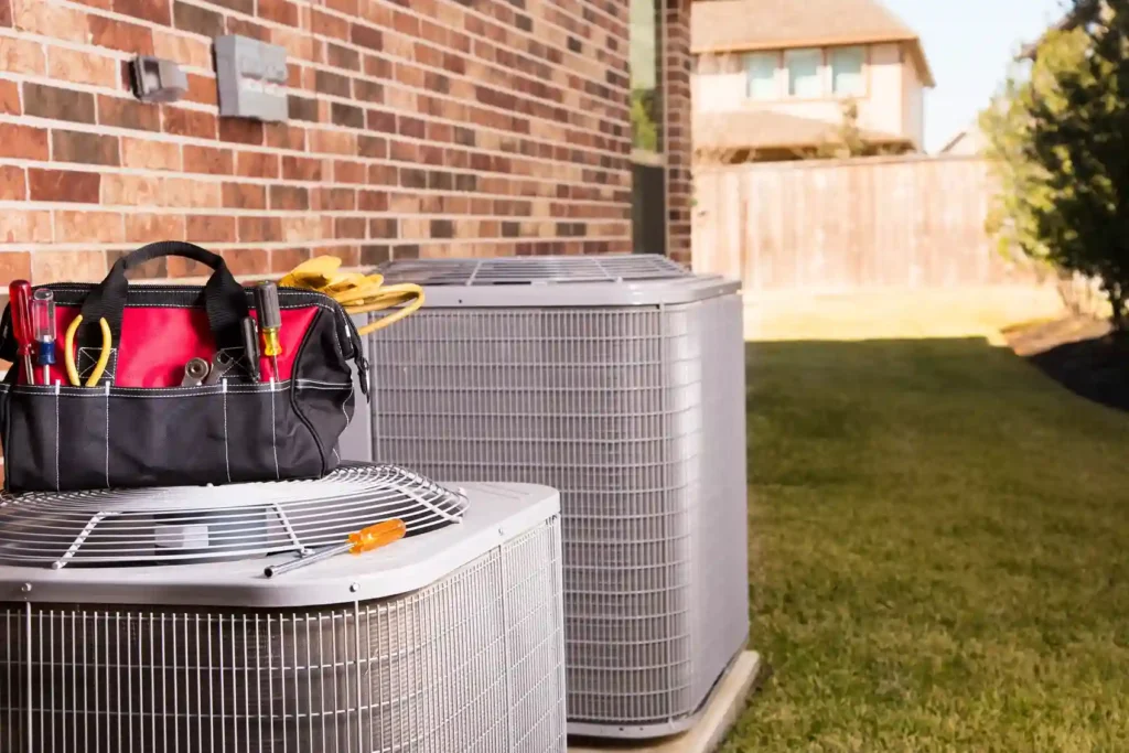 Tool bag with HVAC tools placed on an outdoor air conditioning unit beside a brick house, with a second unit and residential backyard in view