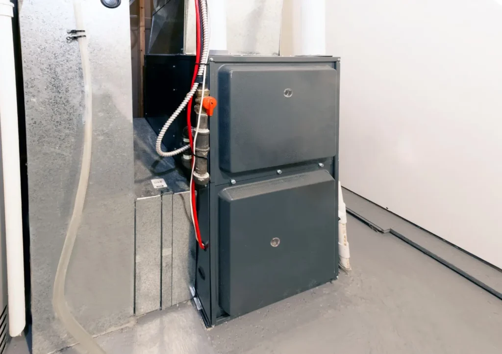 A gray Goodman gas furnace, possibly a GFX10000 model, is installed in a utility room with ductwork and piping visible.