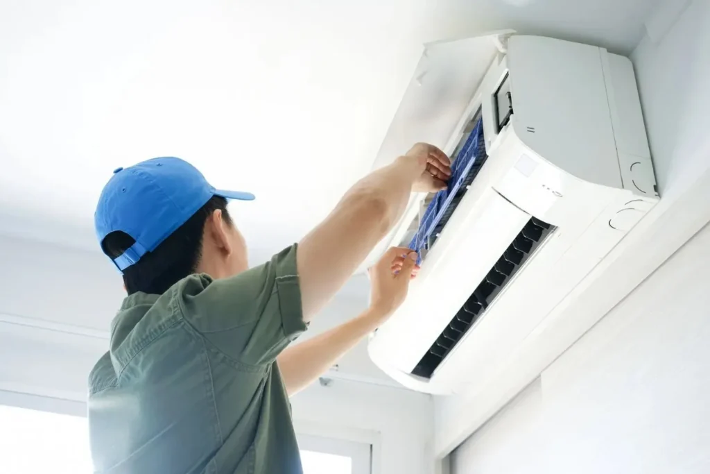 A person wearing a blue cap and a green shirt is shown cleaning or replacing the blue air filters of a white wall-mounted air conditioning unit.